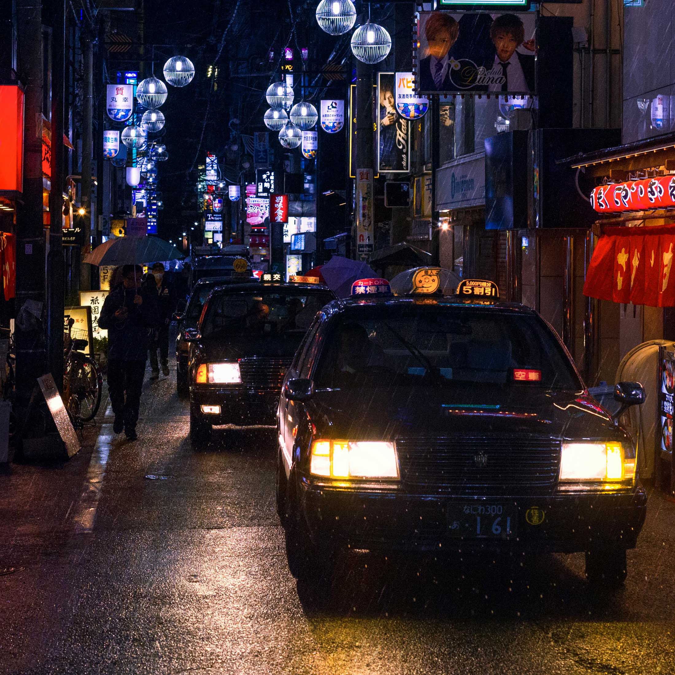 A rainy urban street at night with taxis lined up, their headlights reflecting off the wet pavement.