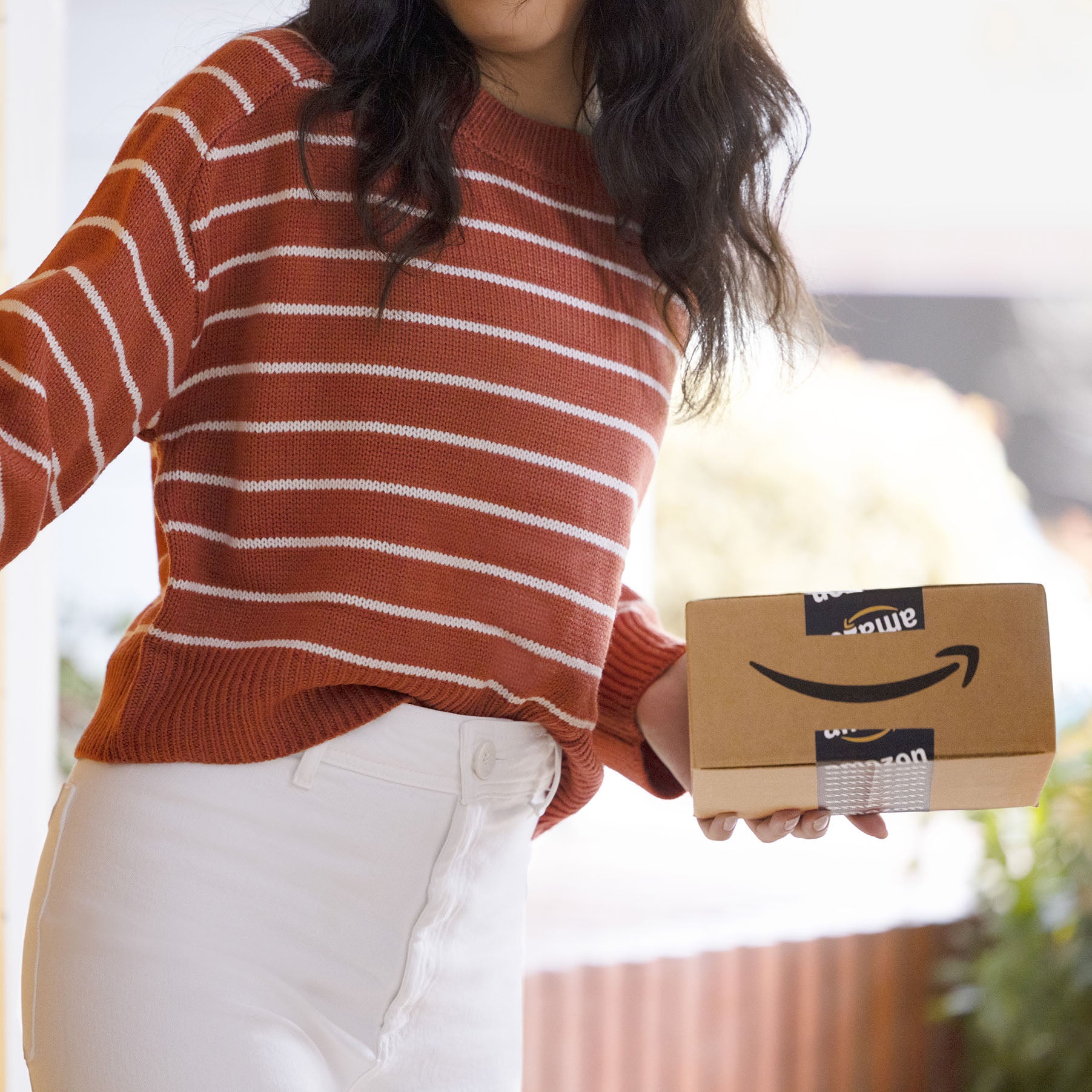 A person wearing a red and white striped sweater and white pants holds a small Amazon delivery box. 