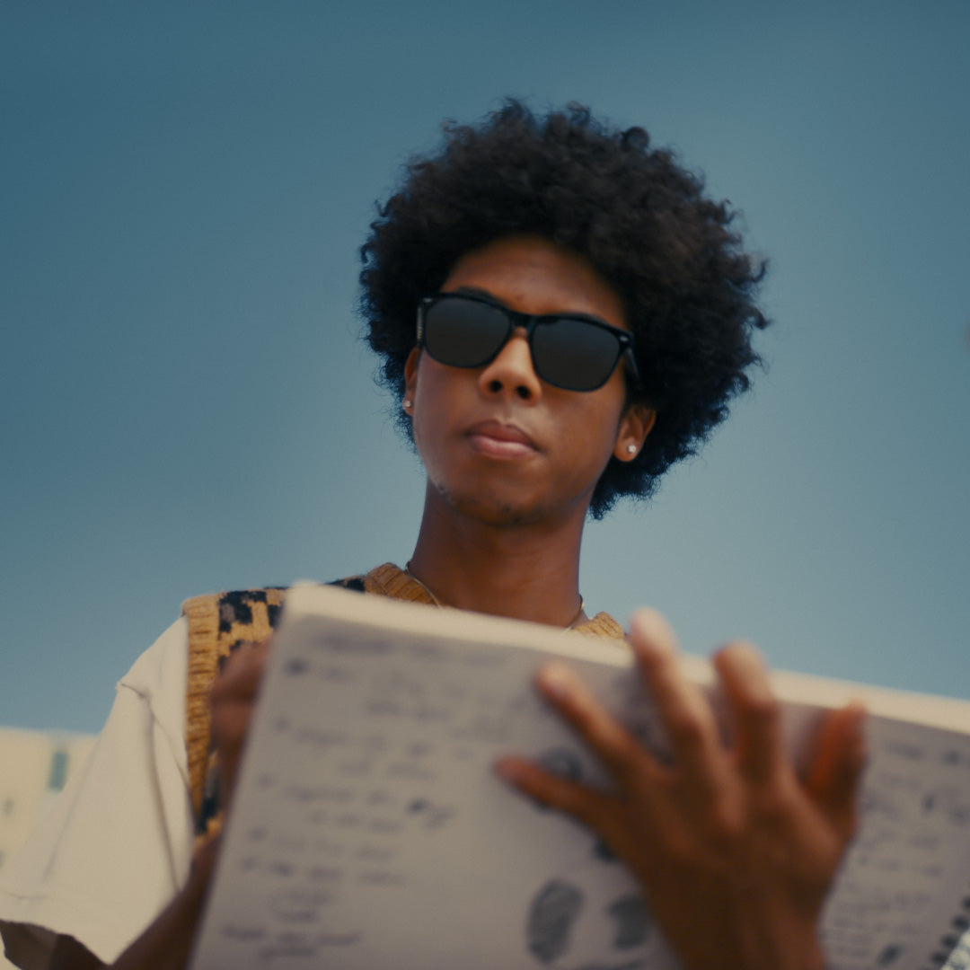 A person with curly hair and wearing sunglasses stands outdoors, holding a notebook filled with sketches or notes.