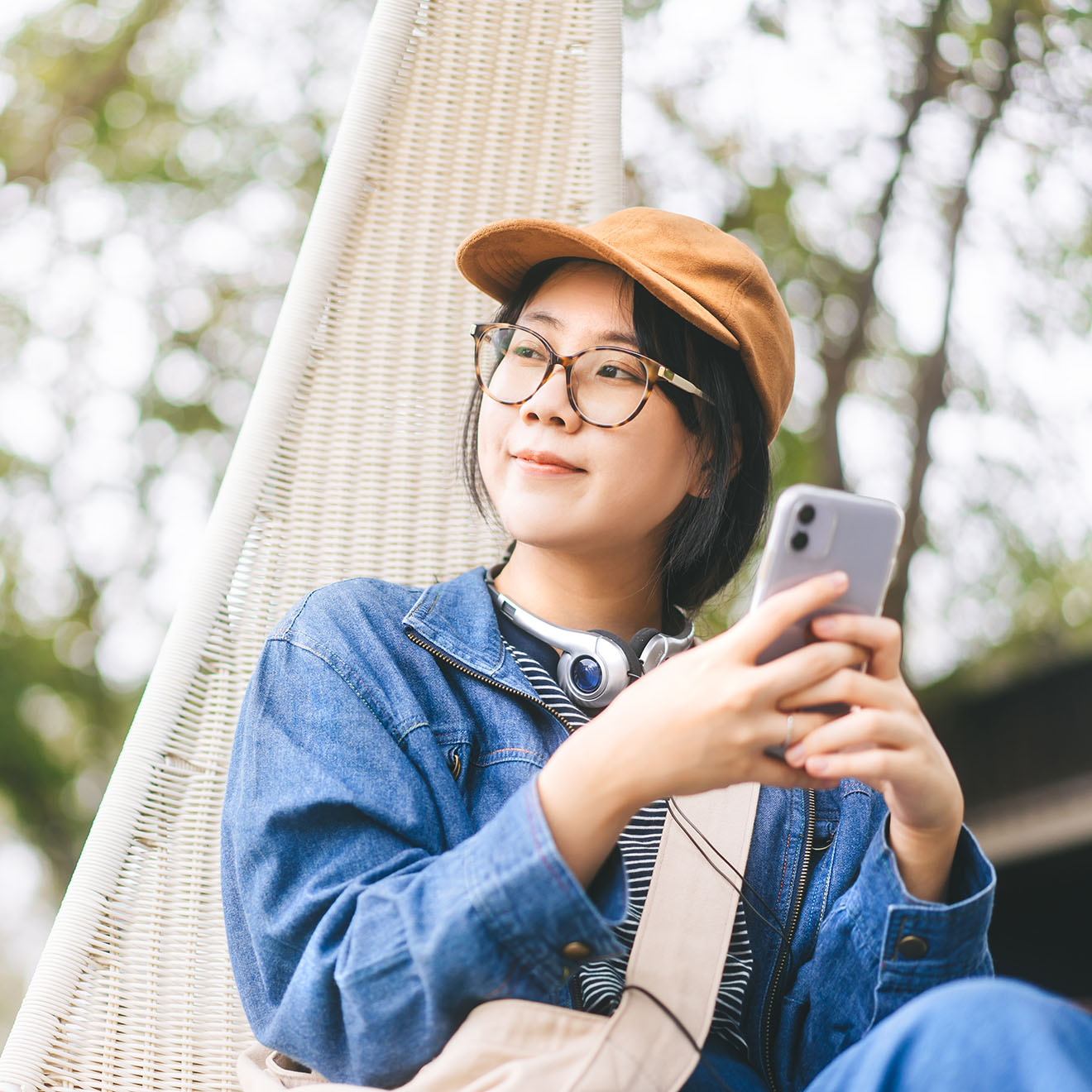 A woman sitting outdoors on a hanging chair, wearing a cap, glasses, and headphones around her neck. She is holding a smartphone and gazing thoughtfully into the distance, dressed casually in a denim jacket.